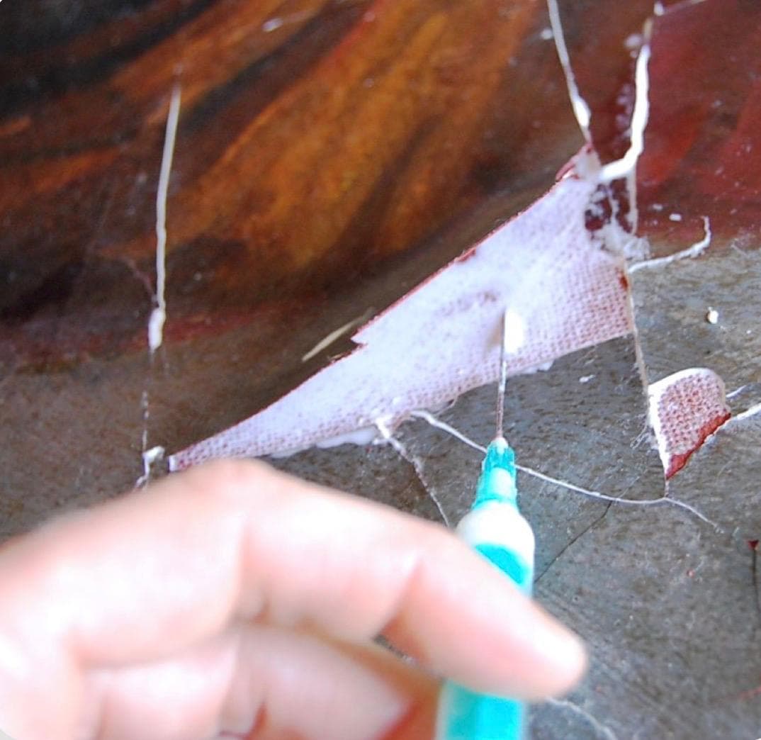 A running crack along a painting's surface, mid-repair with an adhesive syringe