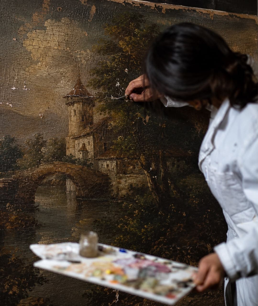 A conservator retouching a landscape oil painting by hand, palette in hand