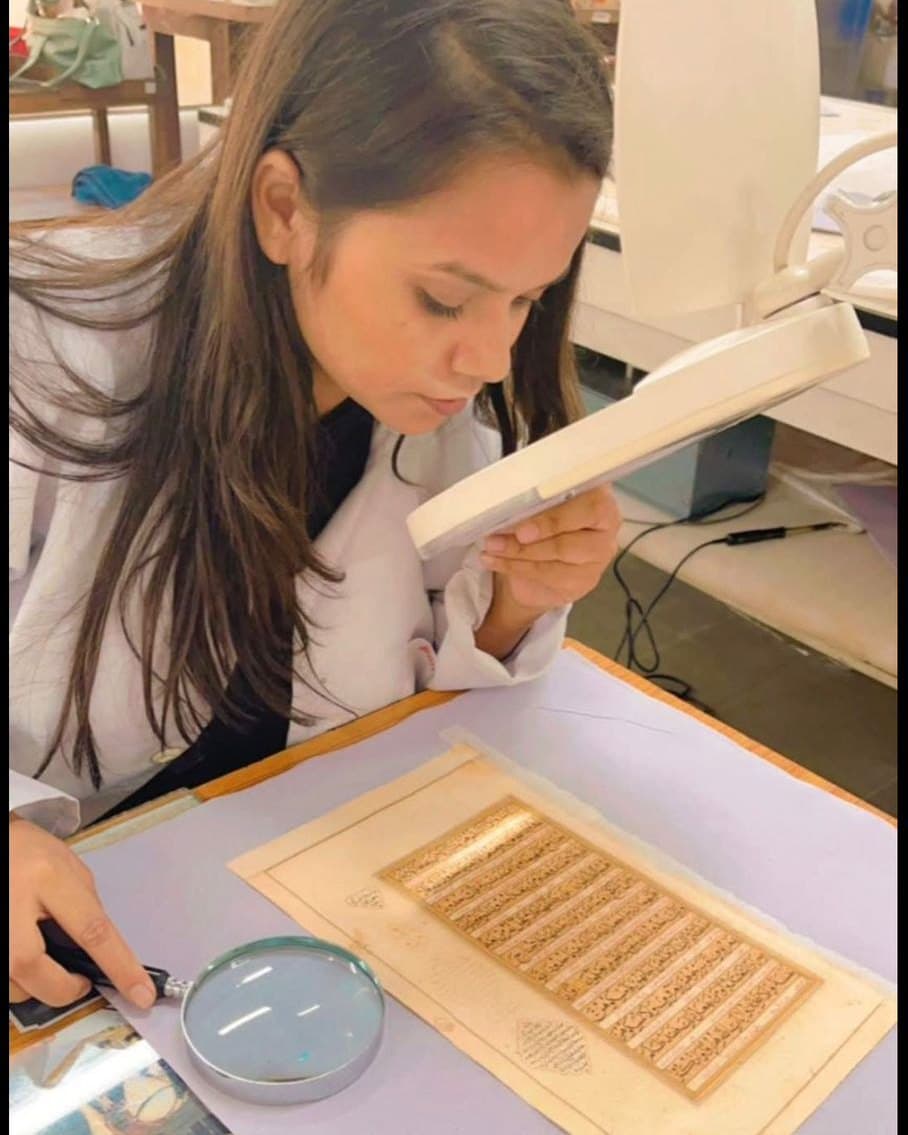 Smriti Rajput examining a manuscript folio under a magnifier and hand loupe in the conservation studio